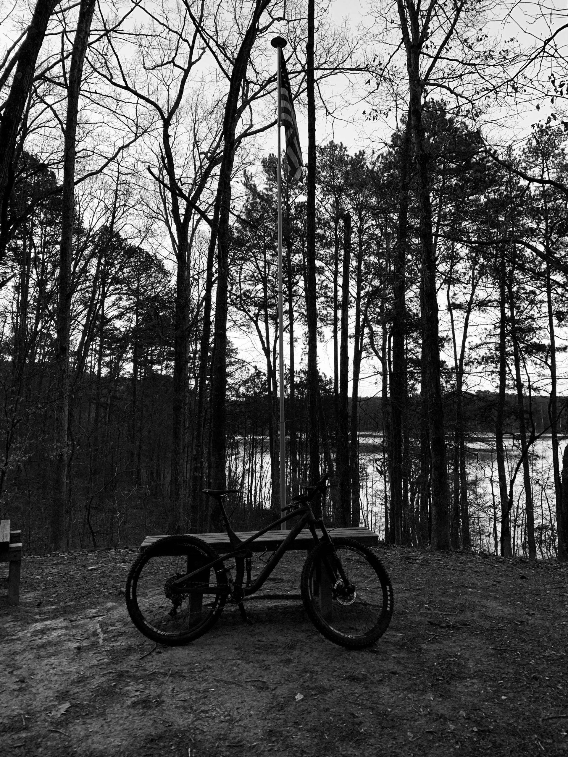A black and white image of a mountain bike resting on a dirt path surrounded by tall trees. In the background, a flagpole with an American flag stands among the trees, and a serene lake can be seen in the distance. There are wooden benches nearby, creating a peaceful outdoor scene. Blankets Creek mountain bike trail.