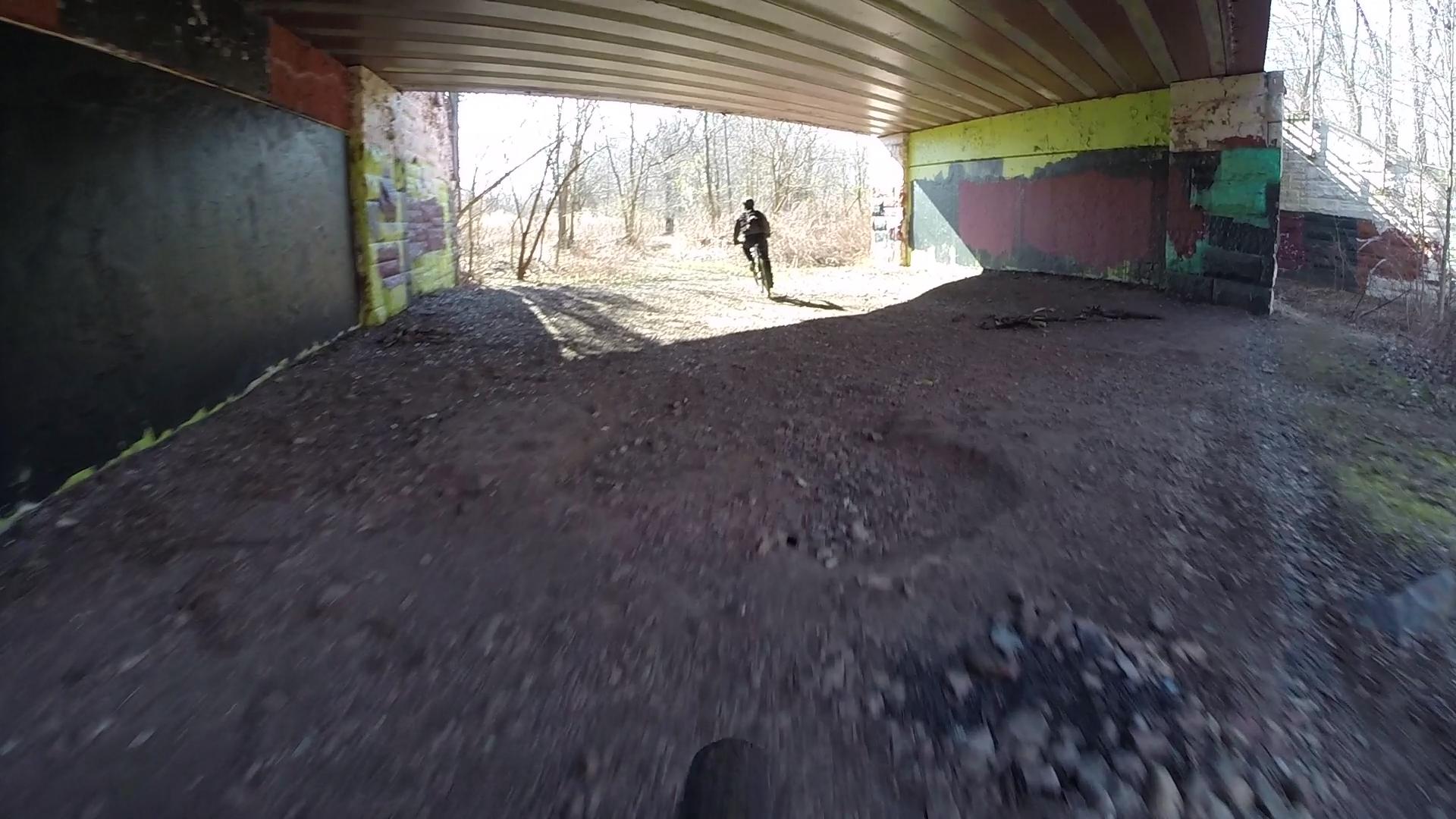 A person riding a bicycle underneath a bridge, with colorful graffiti on the concrete walls. The area is well-lit by sunlight, and trees and brush are visible in the background. The ground is uneven, consisting of dirt and small stones. Richmond Avenue and Forest Hill road mountain bike trail.
