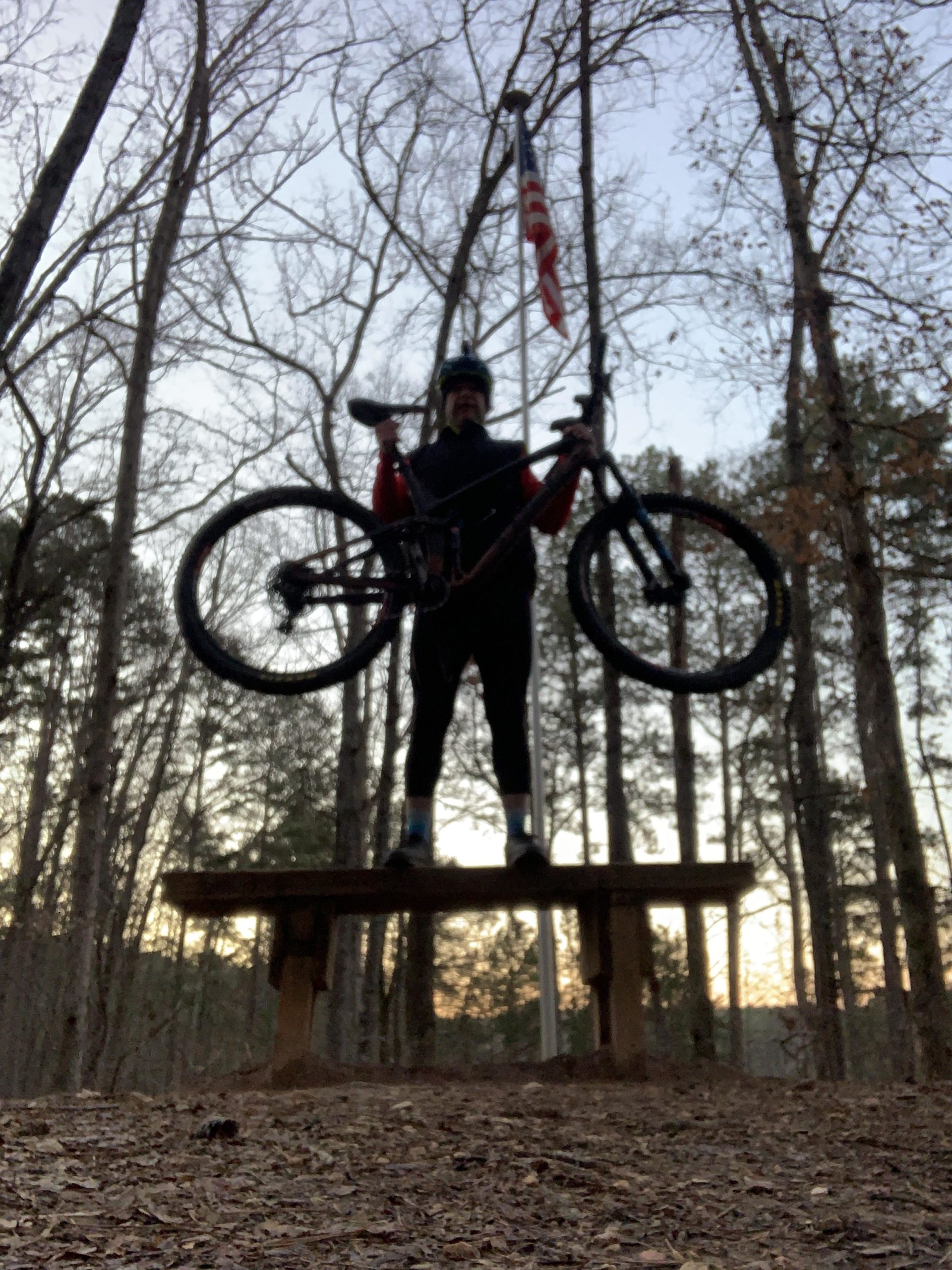 A mountain biker stands triumphantly on a wooden bench in a wooded area, holding their bike above their head. In the background, trees are silhouetted against a twilight sky, and an American flag is visible to the side. The scene captures a moment of celebration and outdoor adventure. Blankets Creek mountain bike trail.