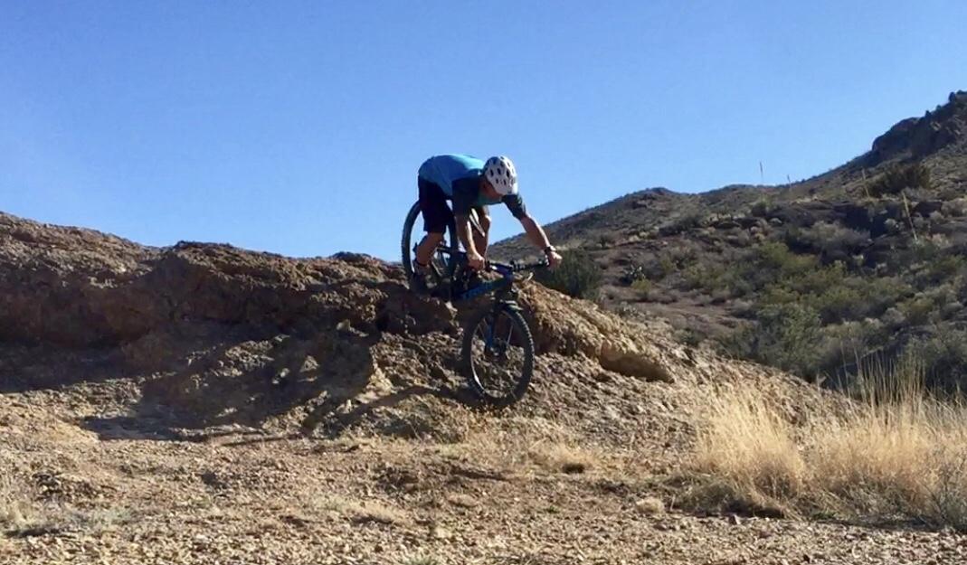A cyclist performing a jumping stunt on a mountain bike over a rocky terrain, with hills and clear blue skies in the background. Franklin Mountain State Park mountain bike trail.