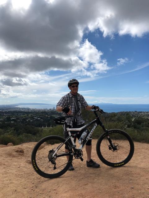 Cannondale Scalpel: A person wearing a helmet and a plaid shirt stands next to a mountain bike on a dirt trail, with a scenic view of hills and the ocean in the background. The sky features a mix of clouds and blue.