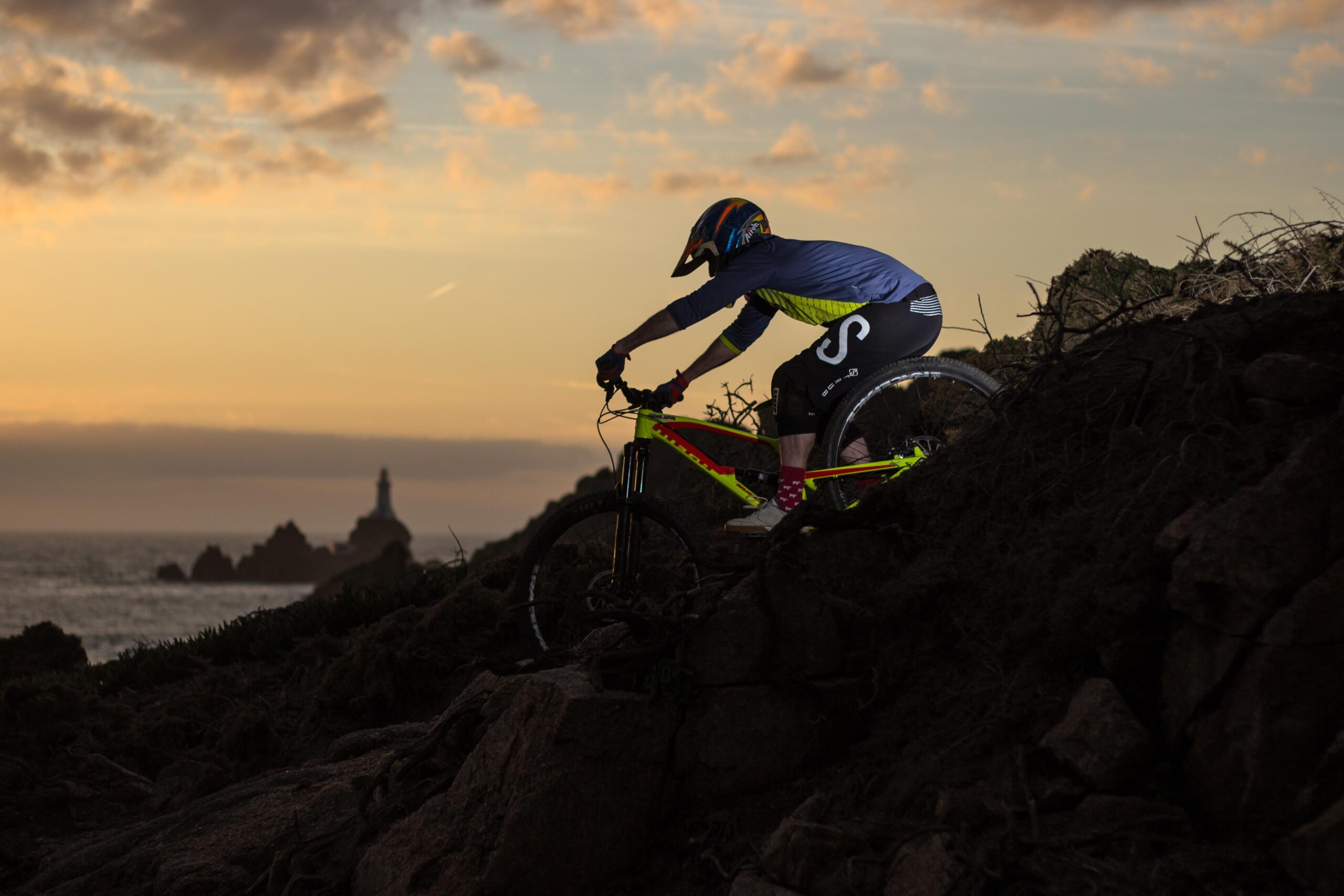 Nukeproof Mega: A mountain biker navigating rocky terrain at sunset, with a distant lighthouse visible on the coastline. The sky features soft clouds illuminated by warm hues, creating a dramatic backdrop. The biker is dressed in a helmet and colorful gear, leaning into the bike as he descends the hill.