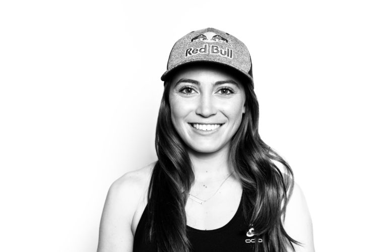 A smiling person wearing a black tank top and a gray cap with a Red Bull logo, posed against a white background.