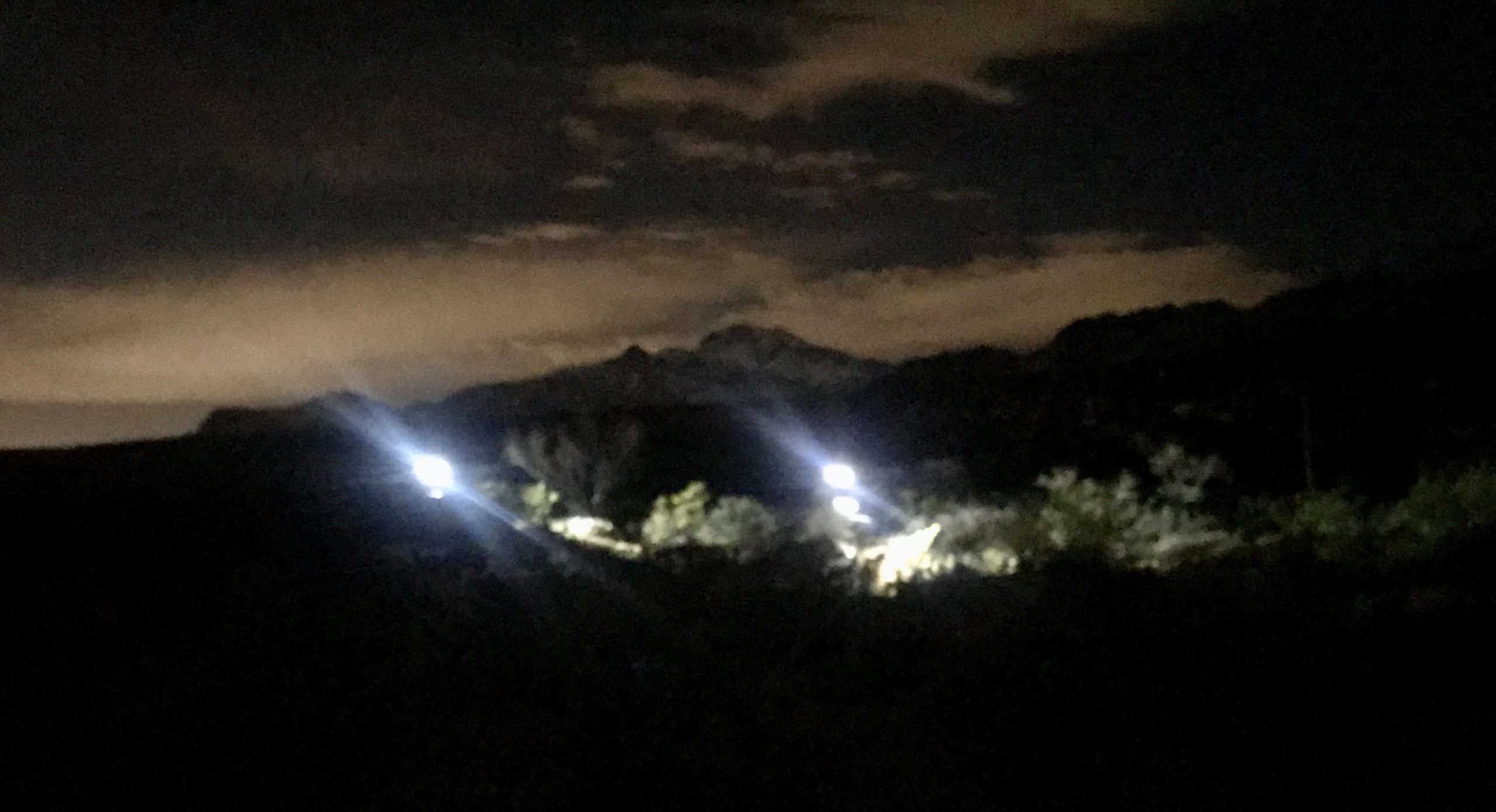 A nighttime landscape featuring distant mountains partially illuminated by light sources, with a cloudy sky above. Franklin Mountain State Park mountain bike trail.