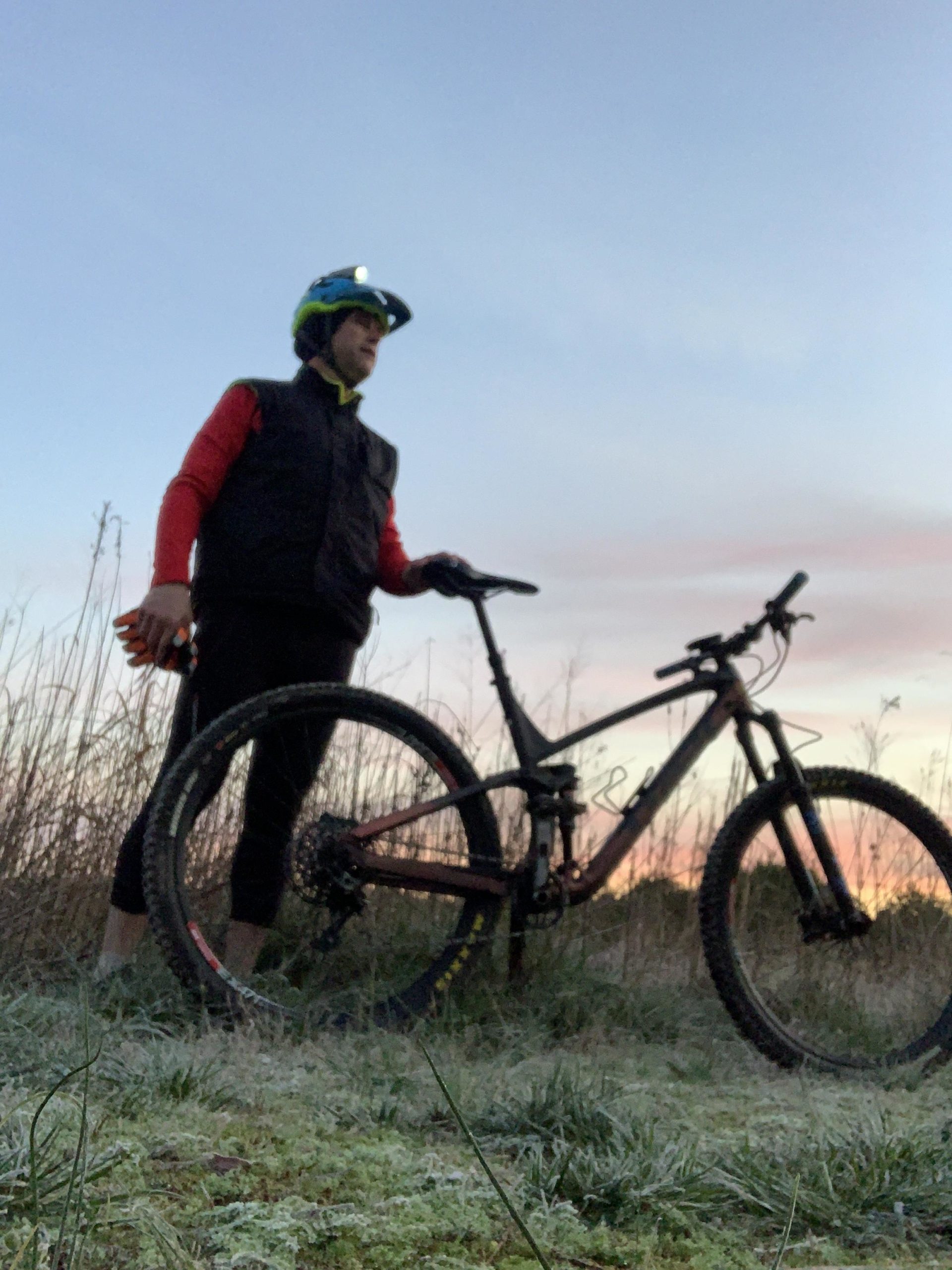 A cyclist wearing a helmet and a vest stands beside a mountain bike in a grassy area at dawn, with a soft, colorful sky in the background. The ground is slightly frosted, indicating early morning temperatures. The cyclist is holding a bike pump in one hand and a water bottle in the other. Allatoona Creek Park mountain bike trail.