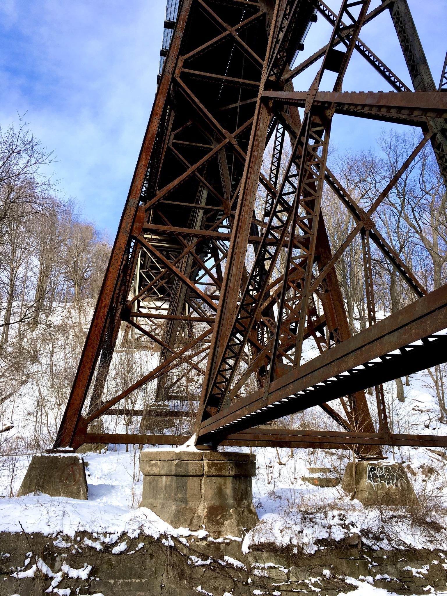 An industrial bridge with a rusted metal structure spans across a snow-covered landscape. The photo, taken from below, captures the bridge's intricate truss design against a backdrop of bare trees and a cloudy blue sky. Snow blankets the ground below the bridge, highlighting the seasonal scenery. Don Valley mountain bike trail.