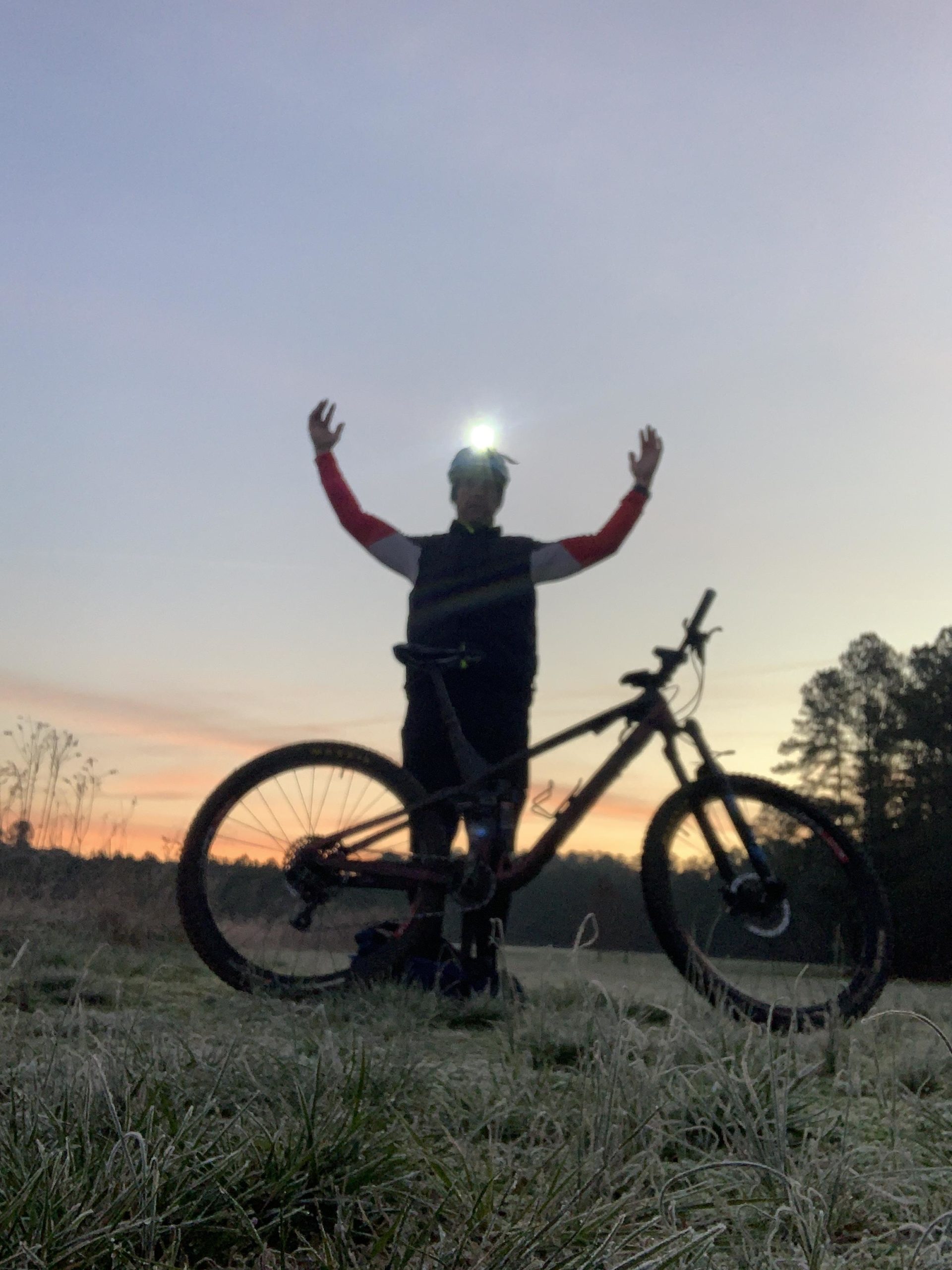 A person wearing a headlamp is standing with arms raised in celebration beside a mountain bike in a grassy field at dawn. The background features a colorful sky with hints of sunrise, and trees are visible in the distance. The ground is covered in frost, suggesting a chilly morning. Allatoona Creek Park mountain bike trail.