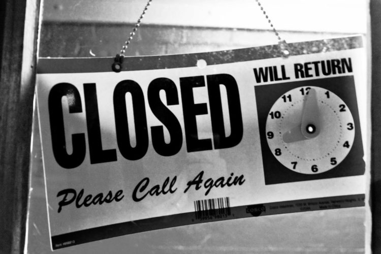 A sign hanging on a door that reads "CLOSED" in large letters, with a clock illustration indicating the time and the message "WILL RETURN" and "Please Call Again" below. The image is in black and white.