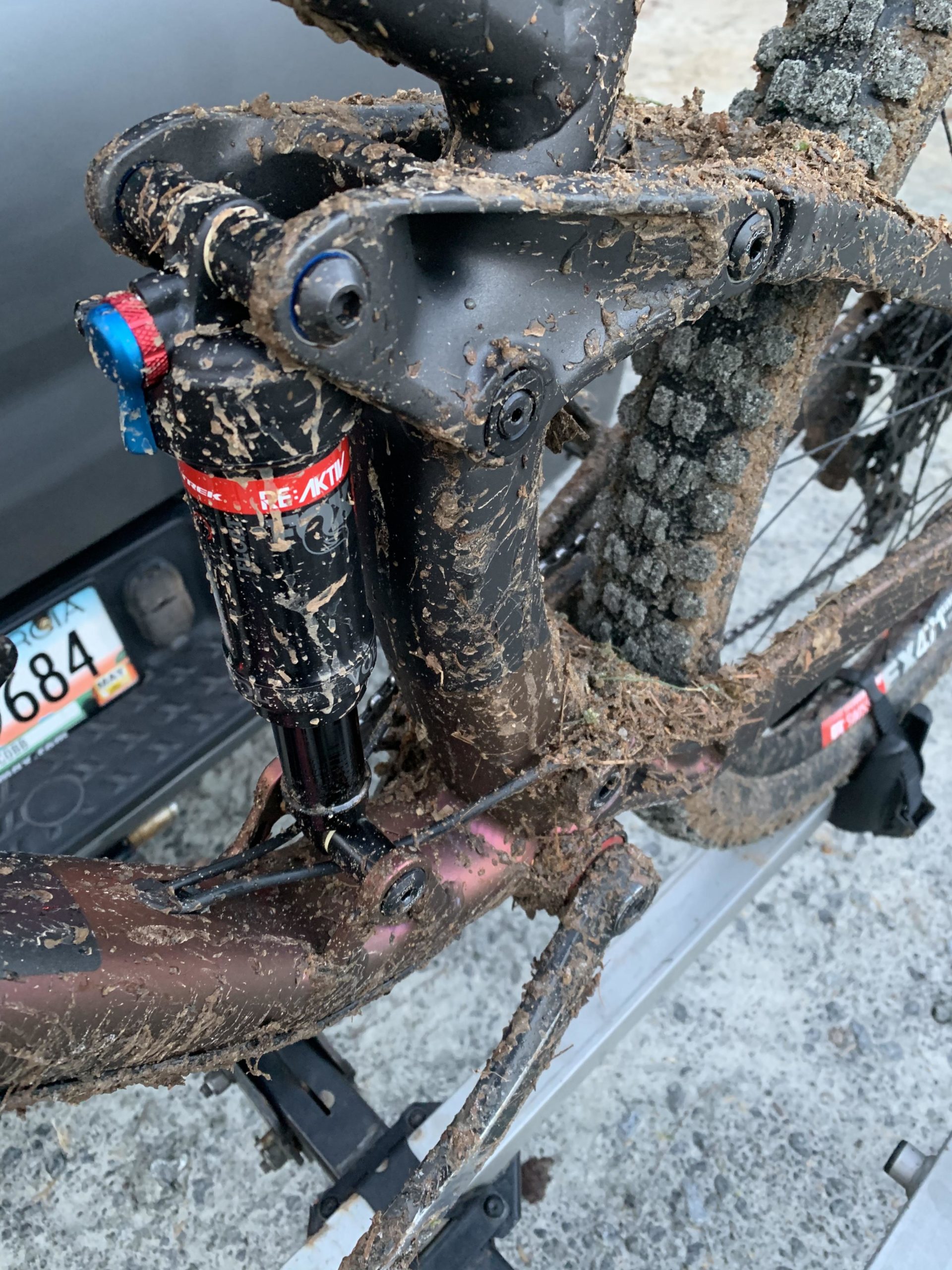Close-up view of a mountain bike's rear suspension and frame covered in mud, highlighting the shock absorber and drivetrain components. The bike is positioned on a rack, with a partially visible license plate in the background. The image captures the details of the muddy condition after a ride. Allatoona Creek Park mountain bike trail.
