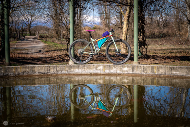 A bicycle is propped up against a green pole beside a still pond, reflecting the bike's colorful gear. In the background, a gravel path meanders through a park, surrounded by bare trees and distant mountains under a clear blue sky.