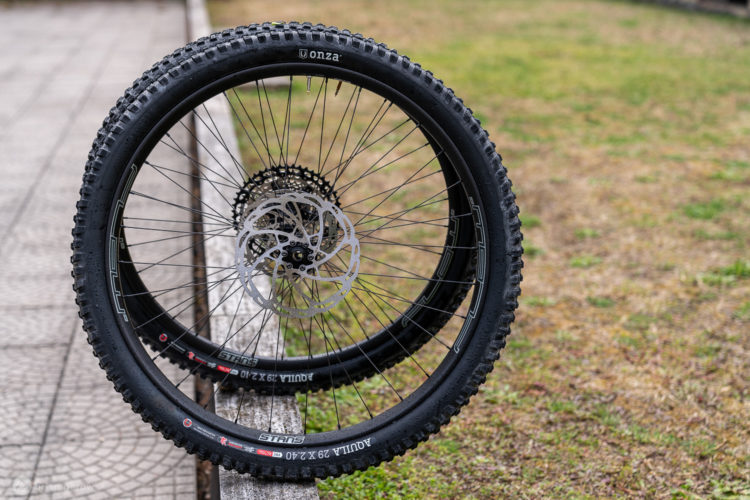 Two mountain bike wheels leaning against a wooden bench, showcasing thick tires and a disc brake rotor. The ground is a mix of grass and pavement, with the wheels appearing shiny, possibly from moisture.