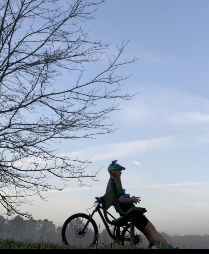 A person sitting on a unique bicycle designed with a chair-like seat, positioned outdoors against a backdrop of a clear sky and a bare tree. The individual is wearing a helmet and appears to be enjoying a serene moment in nature. Allatoona Creek Park mountain bike trail.