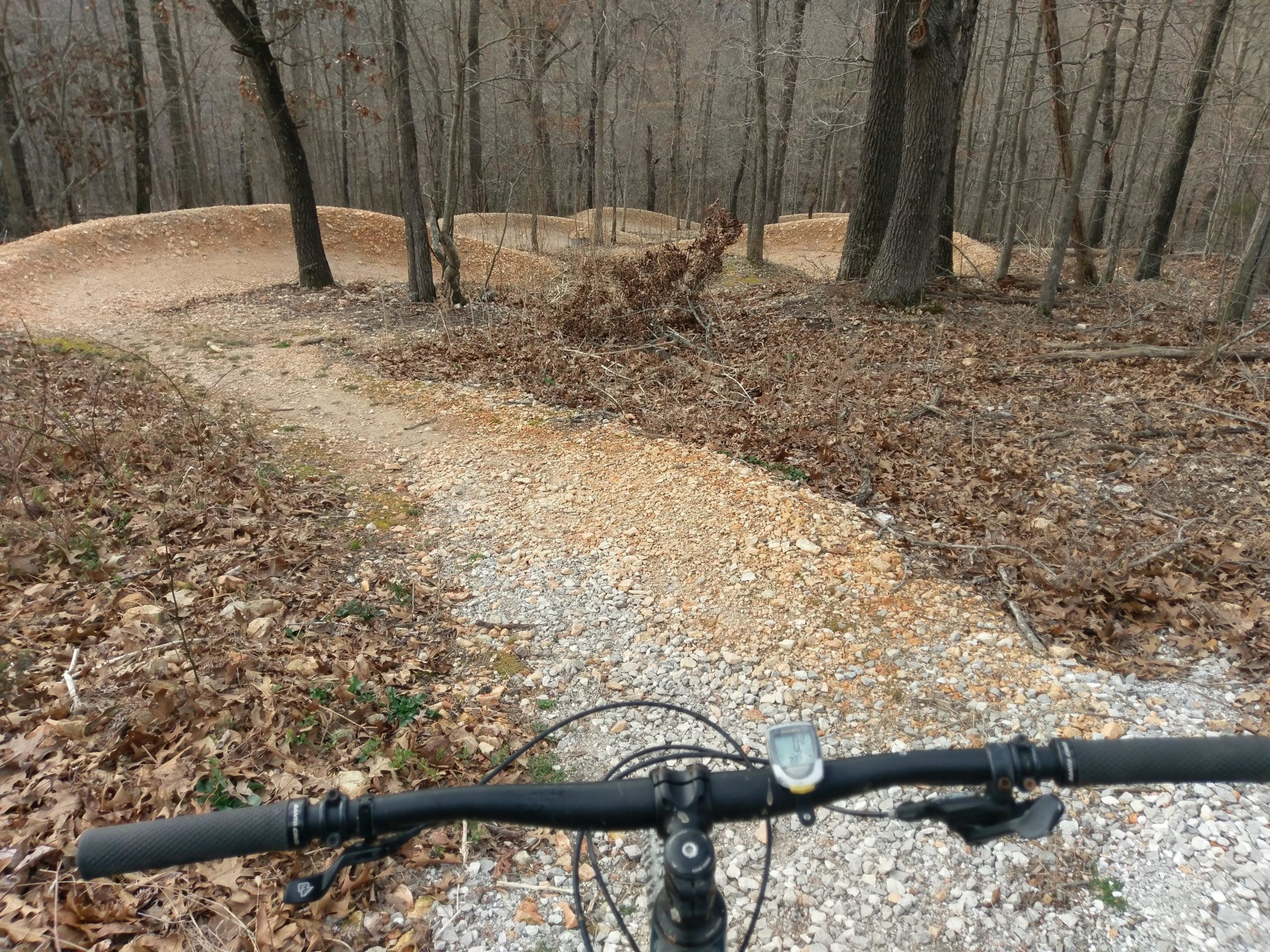 A view from the handlebars of a mountain bike on a winding gravel trail through a forested area, with bare trees and scattered leaves on the ground. The path curves gently ahead, suggesting an adventurous ride through nature. Little Sugar Creek mountain bike trail.