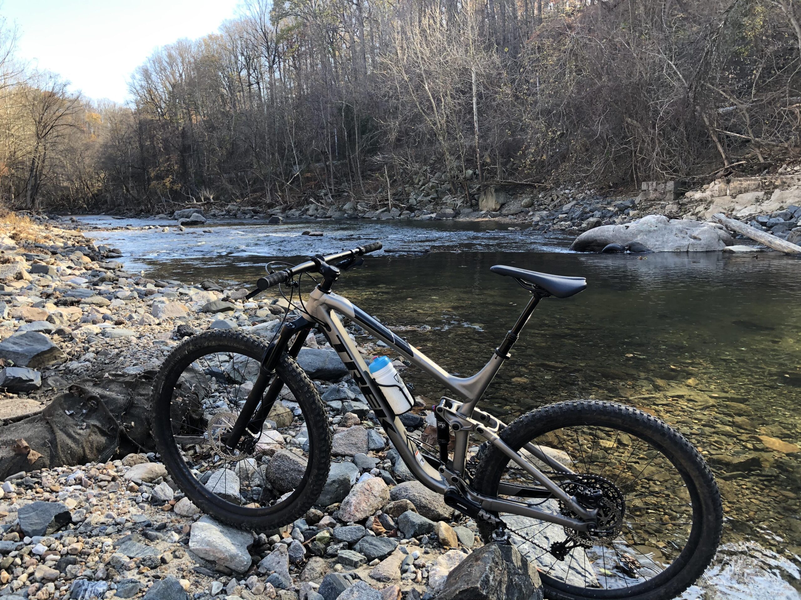 Trek Fuel EX 7 29: A mountain bike resting on rocky terrain beside a gently flowing river, surrounded by sparse trees with autumn foliage. The scene captures a tranquil outdoor setting, inviting exploration and adventure.