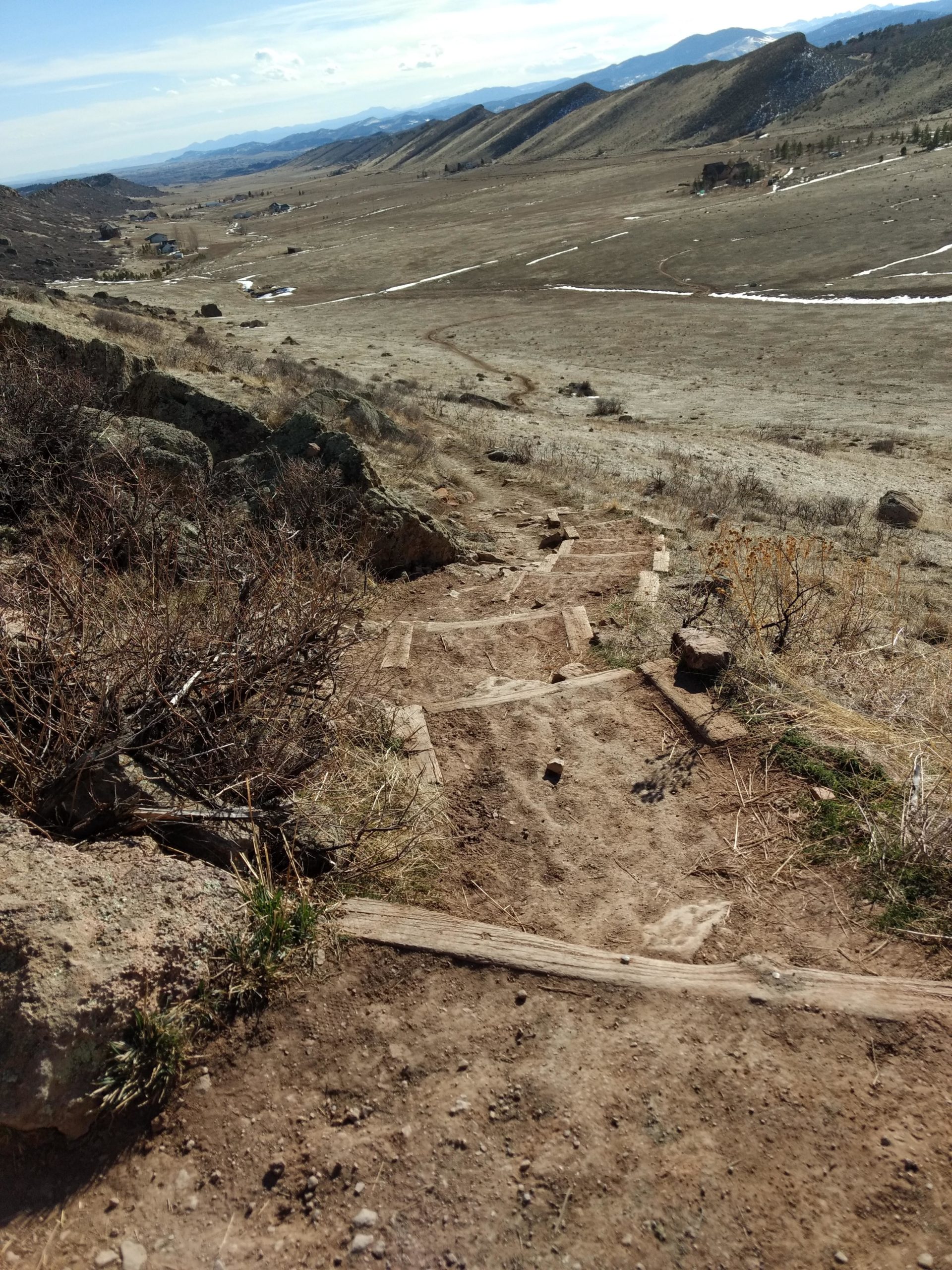 A dirt trail with wooden steps descending through a mountainous landscape. The view includes grassy plains and distant hills under a partially cloudy sky. Coyote Ridge mountain bike trail.