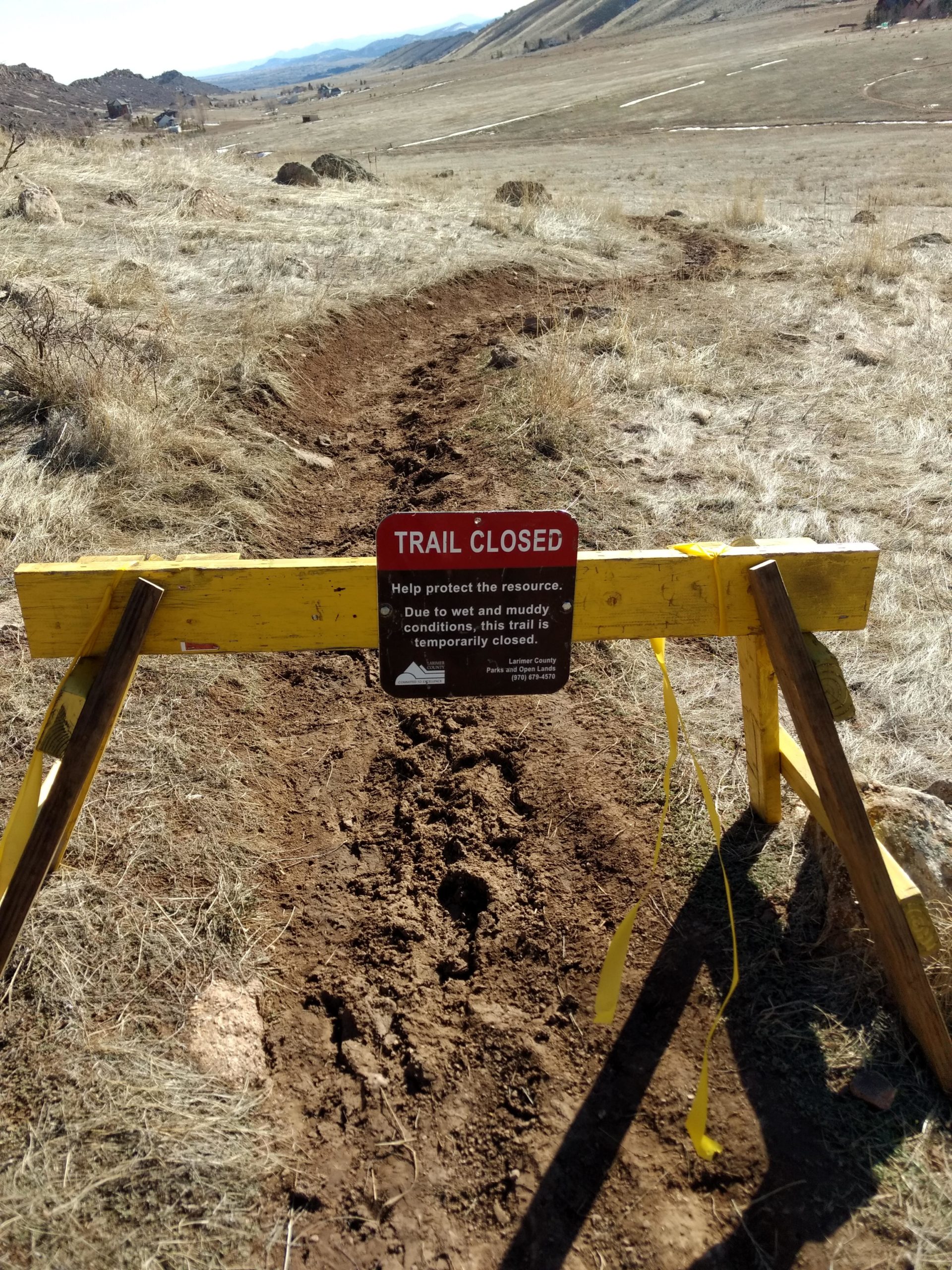 Image of a closed hiking trail, marked by a yellow barrier with a red "TRAIL CLOSED" sign. The sign explains that the trail is temporarily closed due to wet and muddy conditions. The background features a dry, grassy field with some rocky terrain and distant hills. Coyote Ridge mountain bike trail.
