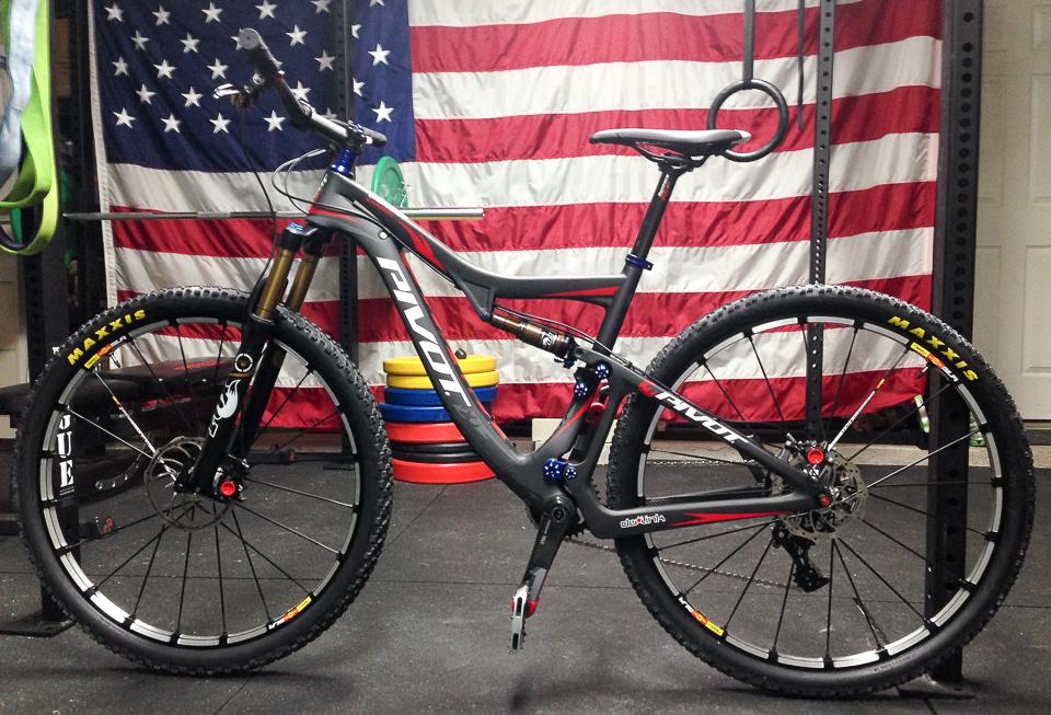 Pivot Mach 429: A black and red mountain bike with the brand name "Pivot" prominently displayed. It features thick tires designed for off-road use, front suspension forks, and a modern frame design. The bike is positioned in a gym or workout space, with a large American flag hanging in the background and colorful weight plates visible on the floor.
