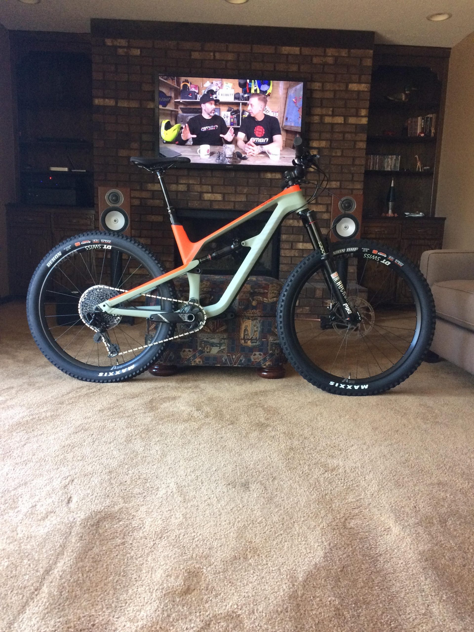 Canyon Spectral CF: A mountain bike with a light blue and orange frame stands on a patterned ottoman in a living room. In the background, a television displays two individuals discussing biking topics, while wooden shelves hold various items. The carpet is a light brown color.