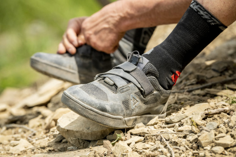 A close-up image of a person fastening a gray sport shoe while sitting on rocky terrain, with a focus on the shoe's details and the surrounding natural environment. The person's leg is partially visible, wearing black socks.