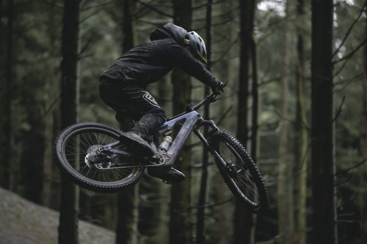 A mountain biker in a black outfit performs a jump over a forested trail, surrounded by tall trees. The rider is in mid-air, showcasing skill and agility while navigating the challenging terrain. The scene is set in a lush, green environment, highlighting the dynamic movement of the sport.