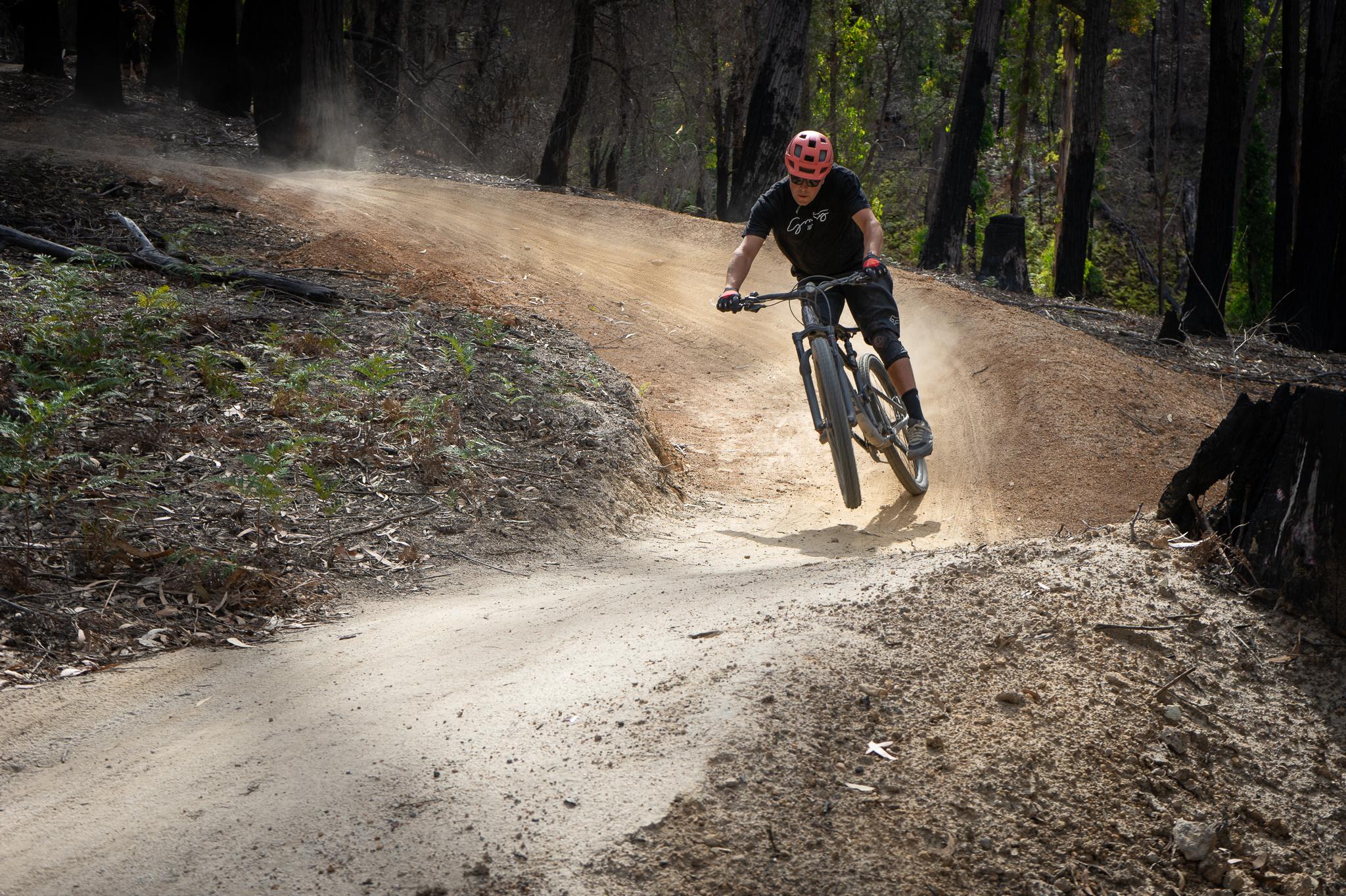 A mountain biker in a pink helmet performs a jump on a dirt trail surrounded by trees and foliage, creating a cloud of dust as they navigate a curved path. St Helens mountain bike trail.