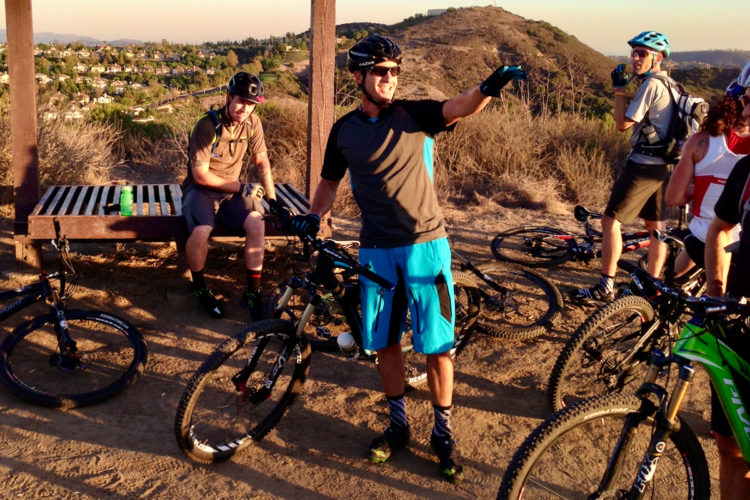 Group of mountain bikers taking a break on a trail. One biker, wearing a helmet and a gray shirt with blue shorts, is smiling and pointing. Another biker is seated on a wooden bench, while others are standing with their bikes nearby. The background features hills and a residential area with greenery. The lighting suggests a late afternoon setting.