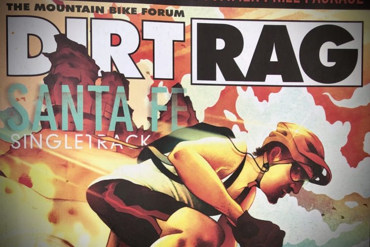 A close-up of a mountain biking magazine cover featuring the title "Dirt Rag." The illustration showcases a cyclist wearing a helmet and sunglasses, racing on a dirt trail with a backdrop of rocky terrain and colorful abstract elements. The words "Santa Fe Singletrack" are prominently displayed below the title.