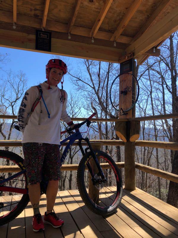 A person wearing a red helmet and casual biking attire stands beside a mountain bike on a wooden platform surrounded by trees. The background features a clear blue sky and distant mountains. A sign is visible on the wall, adding context to the outdoor setting. Fire Mountain Trail System mountain bike trail.
