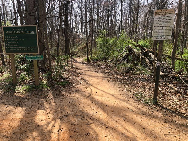 A dirt path leading into a wooded area, bordered by trees and underbrush. A sign on the left welcomes visitors to the mountain bike trail, indicating it is suitable for biking and includes various trail guidelines. Another sign stands nearby with additional information about the trail and its regulations. The scene is illuminated by sunlight filtering through the trees. North Meck Park mountain bike trail.