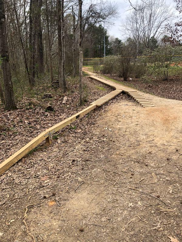 A winding dirt path through a wooded area, featuring a wooden walkway intersecting the trail. The ground is covered with leaves, and trees are visible on either side. North Meck Park mountain bike trail.