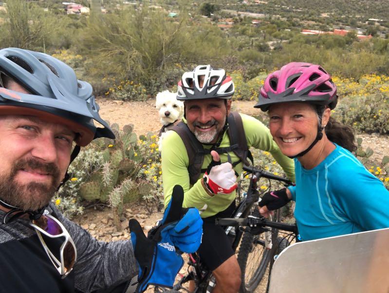 Three mountain bikers posing for a selfie on a trail, surrounded by cacti and wildflowers. Two men and one woman are smiling, wearing helmets and biking gear. One man gives a thumbs-up, while a small dog peeks out from a backpack behind him. The background features a hilly landscape with greenery and a glimpse of a town in the distance. Tucson Mountain Park mountain bike trail.