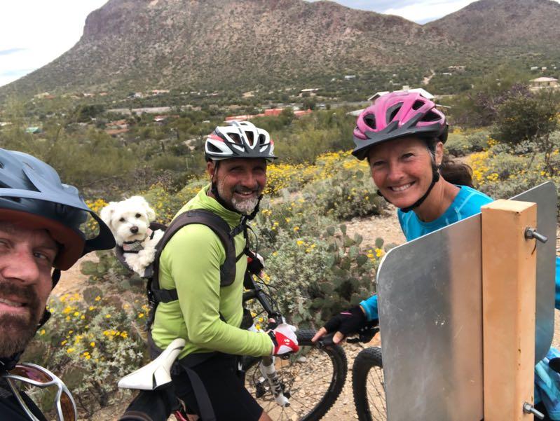 A group of three mountain bikers posing for a selfie on a trail surrounded by desert scenery. One of the bikers has a small dog in a backpack. They are smiling, wearing helmets, and cycling gear, with a mountain in the background and wildflowers in bloom. Robles Pass mountain bike trail.