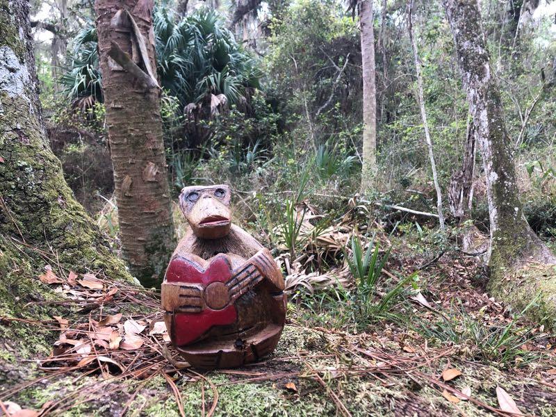 A wooden carved figure of a frog playing a guitar, set against a natural background of trees and lush greenery in a forested area. Pine needles and small plants surround the figure on the ground. Moses Creek mountain bike trail.