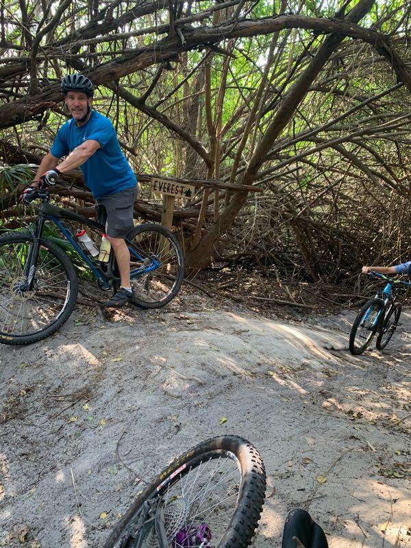 A man wearing a blue shirt and a helmet is smiling while standing beside his bicycle on a sandy trail, with a sign in the background reading "Everest." A child on a blue bike can be seen in the background, surrounded by lush greenery and branches. Fort Pierce Mountain Bike Trail mountain bike trail.