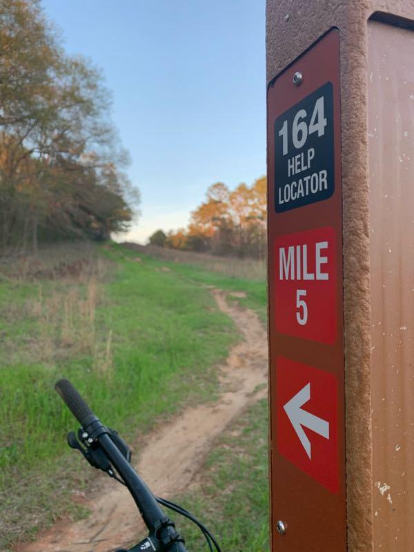 A trail sign indicating a help locator with the number 164 and marking mile 5 on a dirt path. A bicycle is partially visible in the foreground, and the background features a grassy trail leading into a wooded area under a clear sky. Georgia International Horse Park mountain bike trail.