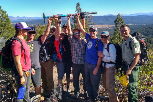 A group of eight people, including one wearing a ranger uniform, pose together on a mountain ridge, smiling and celebrating. One person holds up a tool, while the others cheer and smile at the camera. A black dog is in the foreground, and the scene features trees and a scenic view of mountains in the background under a clear blue sky.