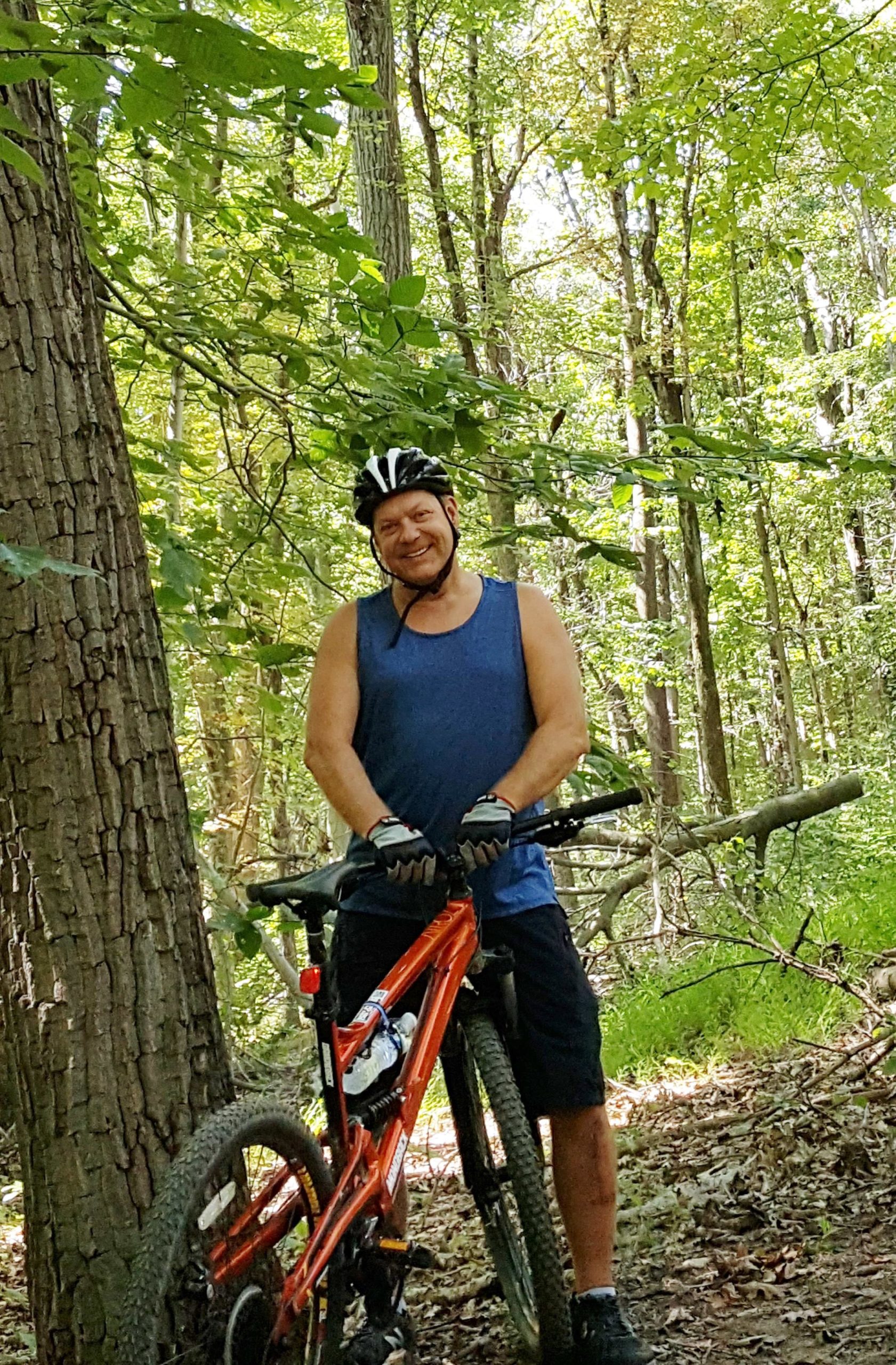 A smiling person in a blue tank top and black shorts stands beside an orange mountain bike, leaning against a tree in a wooded area. The background features dense green foliage and trees, indicating a natural, outdoor setting. The person is wearing a black helmet and gloves, suggesting they are engaged in mountain biking. Lewis Morris mountain bike trail.
