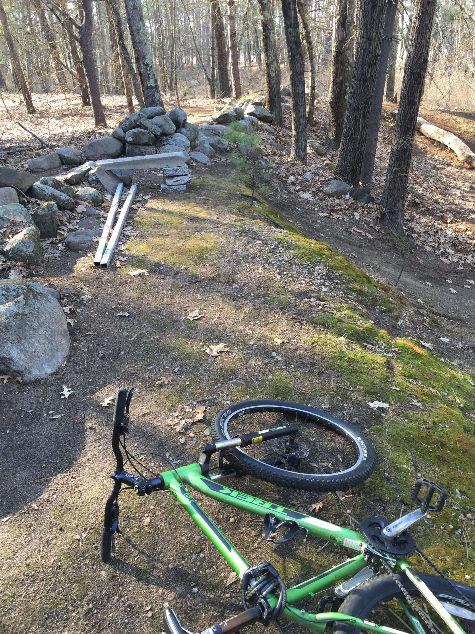 A green mountain bike lies on its side on a dirt path surrounded by trees. Nearby, a stack of rocks and some metal pipes are visible. The ground is covered in moss and scattered leaves, indicating a wooded area during early spring or fall. Hitchinpost Greens mountain bike trail.