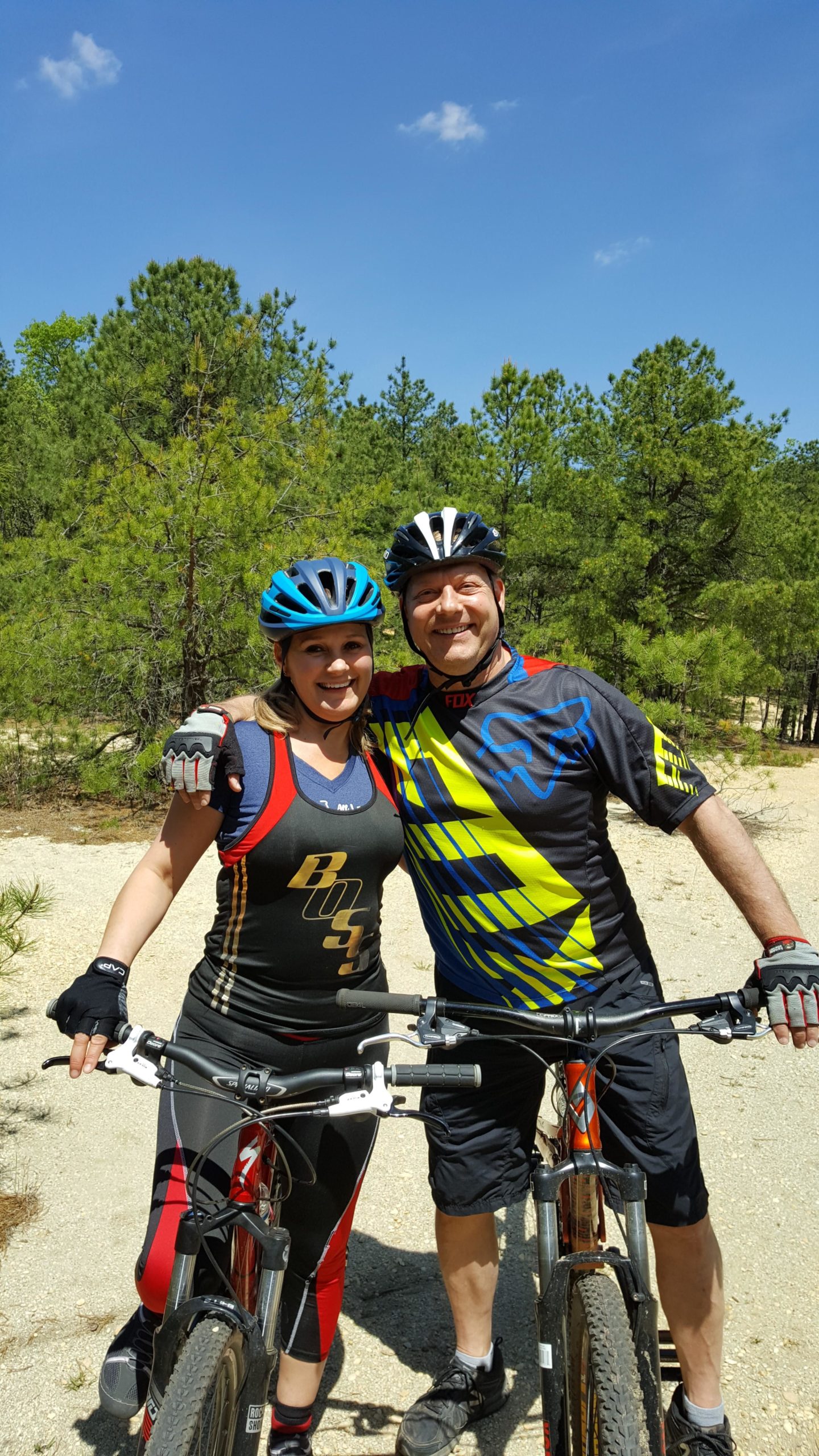 A smiling woman and man pose together on mountain bikes in a sunny outdoor setting, surrounded by green pine trees. The woman is wearing a black tank top with gold lettering, black and red leggings, and a blue helmet. The man is dressed in a colorful blue and yellow jersey with black shorts and a black helmet. They appear happy and ready for cycling. Allaire State Park mountain bike trail.