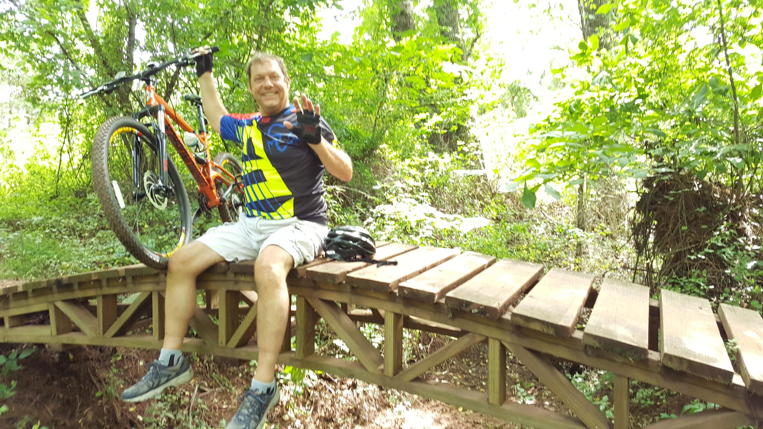 A person sitting on a wooden bridge in a forested area, smiling and waving. They are holding an orange mountain bike and wearing a brightly colored cycling shirt, shorts, and a bicycle helmet. Lush greenery surrounds them, creating a vibrant, outdoor atmosphere. Six Mile Run mountain bike trail.
