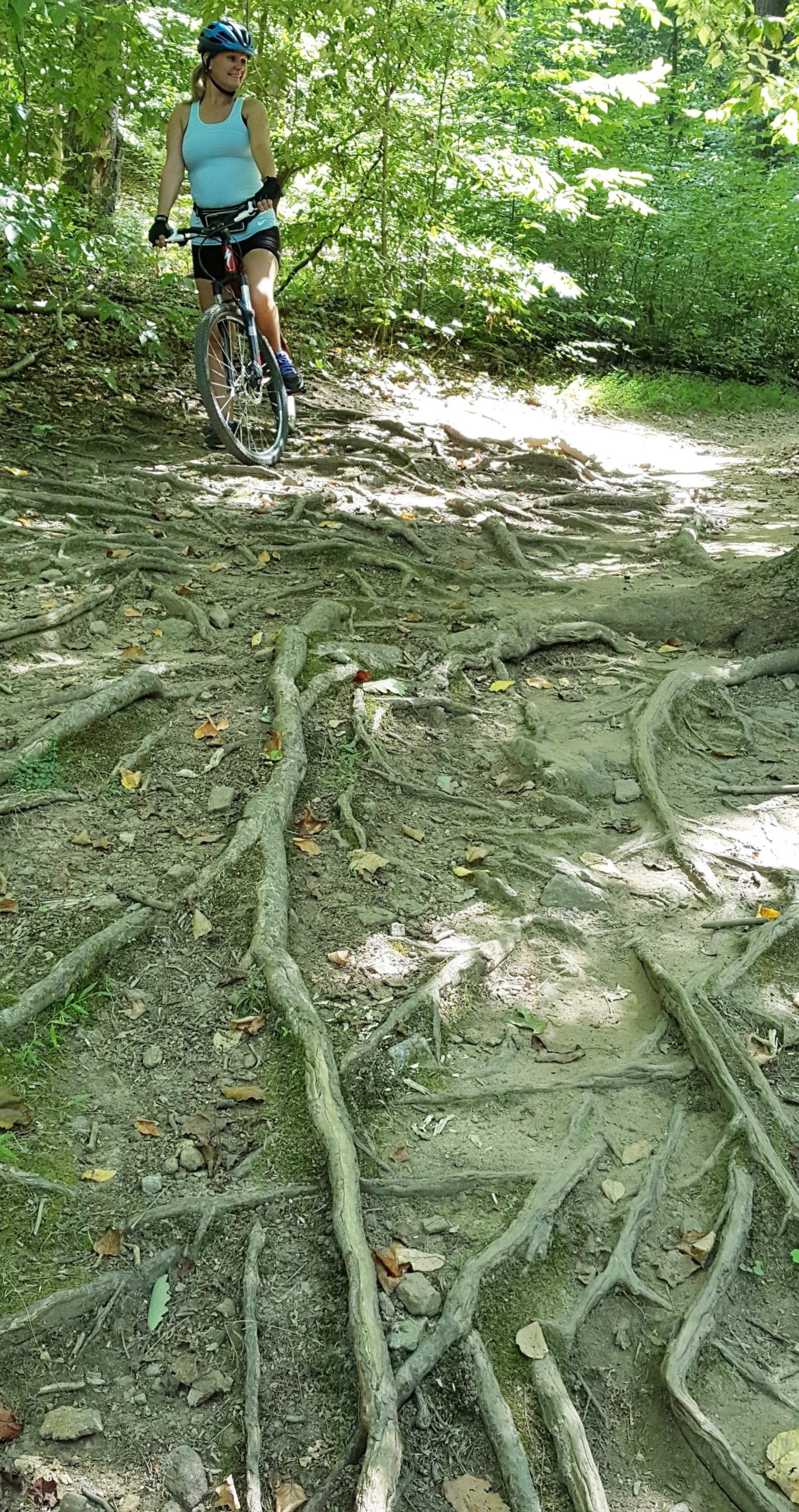 A woman wearing a helmet and athletic attire stands beside her mountain bike on a wooded trail. The ground features exposed tree roots and patches of dirt, surrounded by lush green foliage and dappled sunlight filtering through the trees. Lewis Morris mountain bike trail.