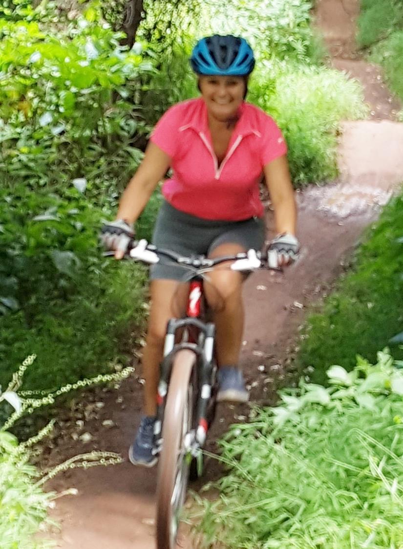 A person riding a mountain bike along a narrow, dirt pathway surrounded by lush greenery. They are wearing a pink jersey, a blue helmet, and shorts, smiling as they navigate the trail. Allaire State Park mountain bike trail.