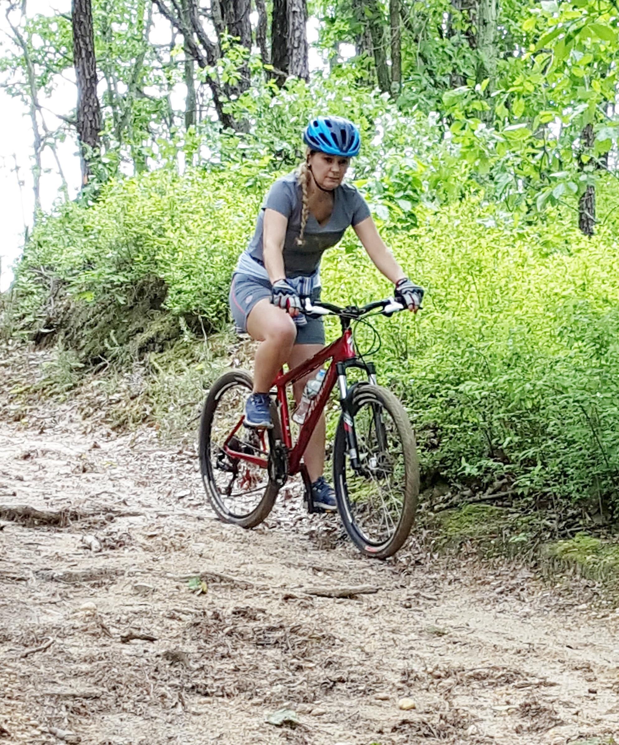 A person riding a red mountain bike along a dirt trail in a wooded area. They are wearing a blue helmet and a gray t-shirt, with their hair braided. Lush green foliage surrounds the path, creating a natural, outdoor setting. Six Mile Run mountain bike trail.