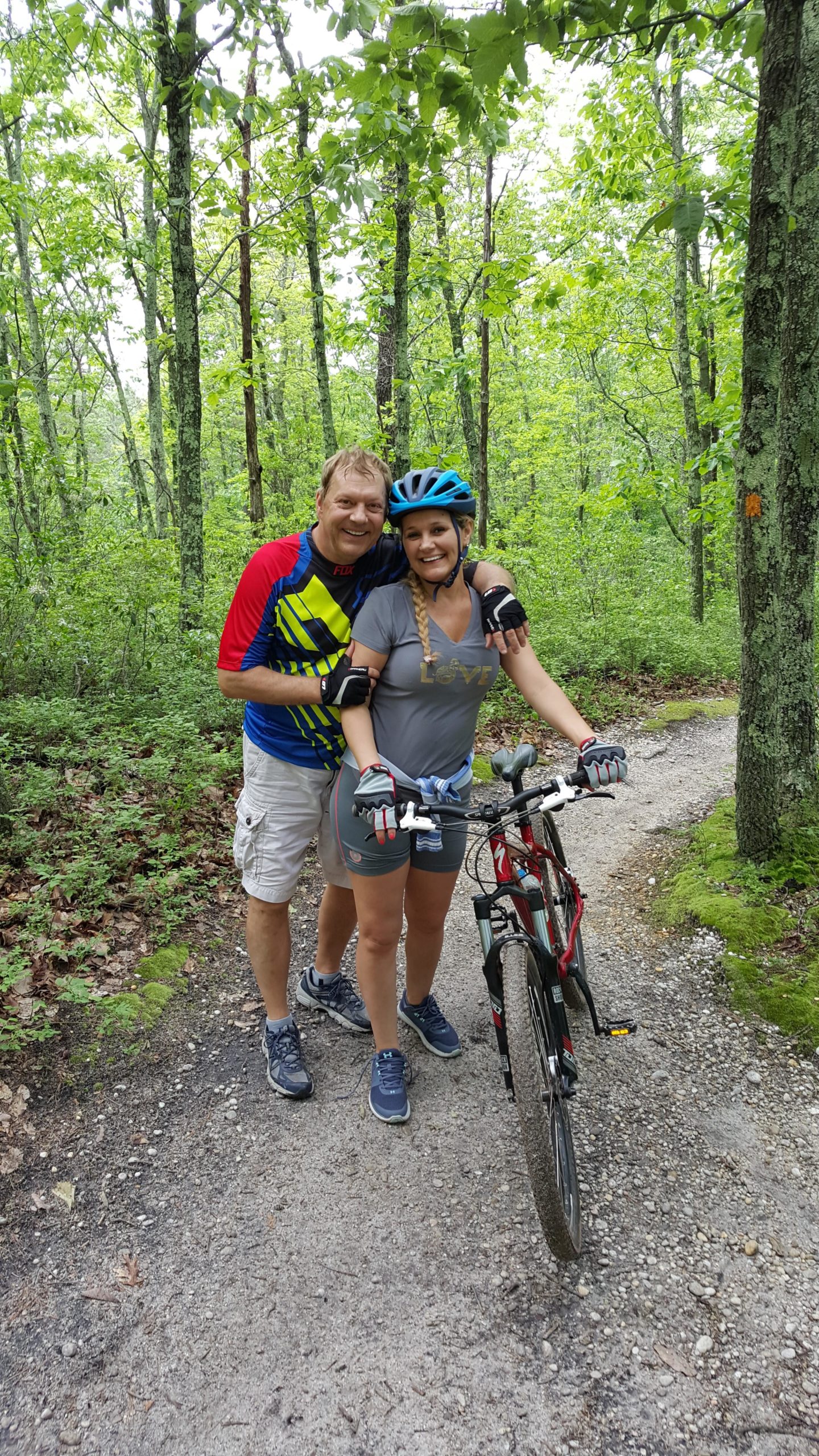 A man and a woman pose together on a forested bike trail, smiling and enjoying their time outdoors. The man is wearing a colorful cycling jersey, while the woman is in a gray shirt with the word "LOVE" printed on it. Both are wearing cycling gloves, and the woman has a blue helmet on. A mountain bike is parked beside them on the gravel path, surrounded by lush green trees and foliage. Six Mile Run mountain bike trail.