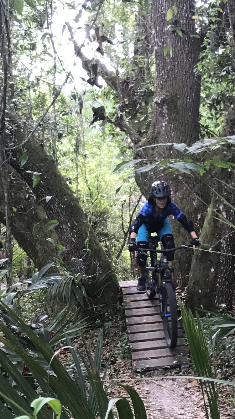 A mountain biker riding over a wooden bridge in a lush green forest. The path is surrounded by tall trees and thick vegetation, creating a natural trail atmosphere. The biker is wearing a helmet and protective gear, focusing intently on navigating the trail. Chuck Lennon Park mountain bike trail.