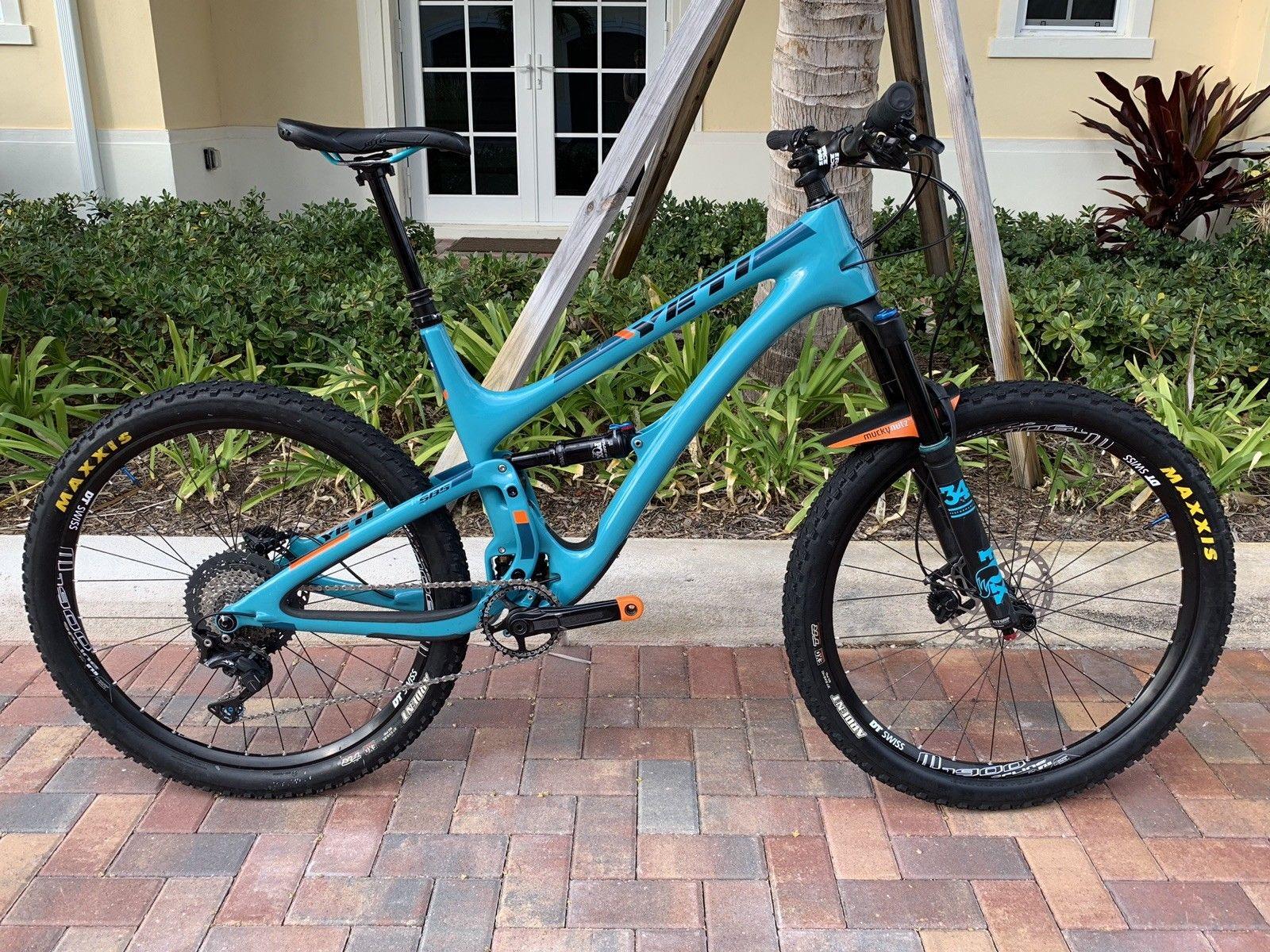 Yeti 2018 Yeti SB5 Mountain Bike Large 27.5 Carbon: A blue mountain bike parked on a paved walkway, featuring thick tires and a sturdy frame. The bike is equipped with a suspension fork, and there are visible components such as gears and a chain. Lush green shrubs and a building can be seen in the background.