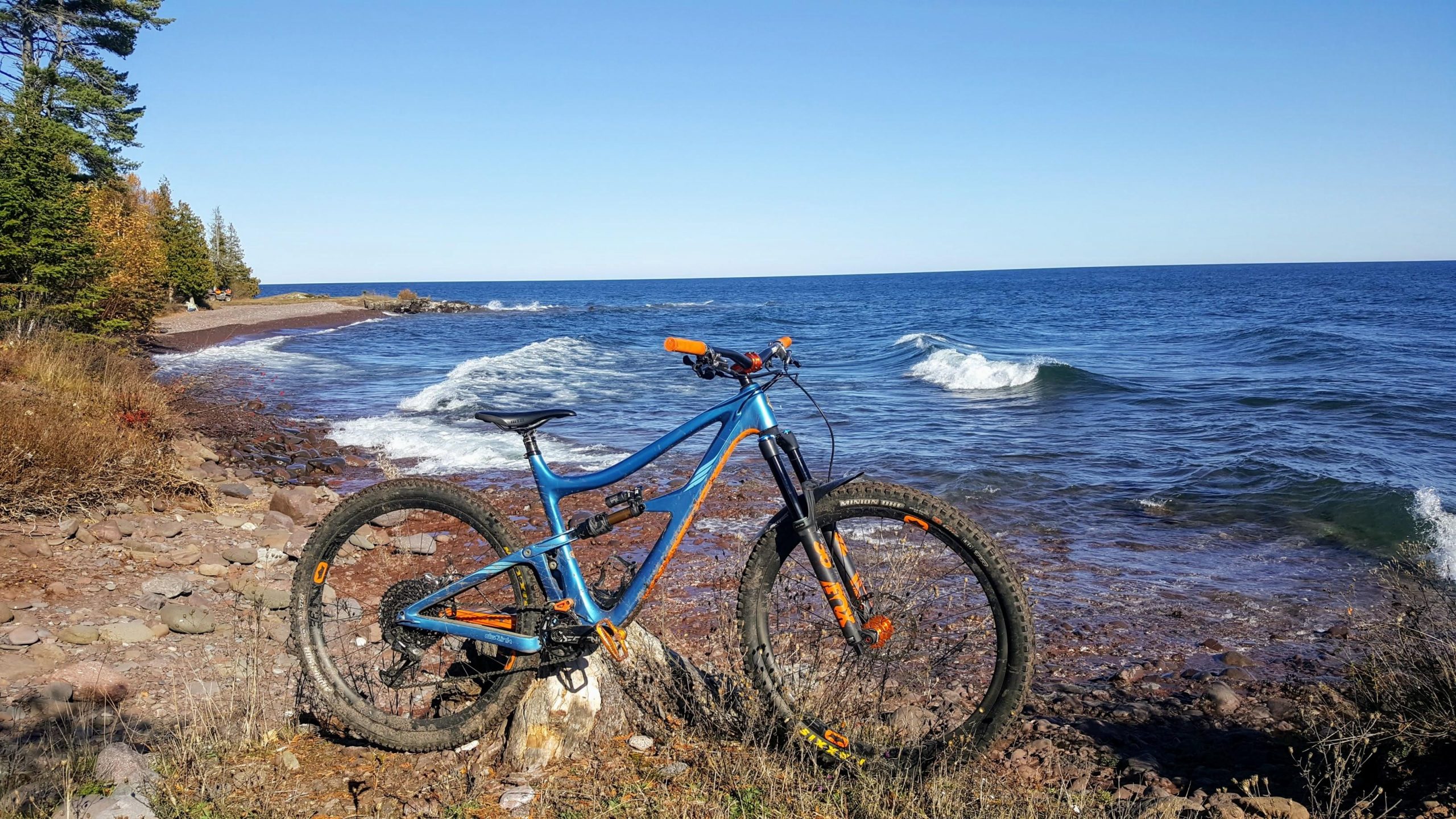 Ibis RipMo: A blue mountain bike with orange accents is parked on a rocky shoreline, overlooking a body of water with gentle waves. In the background, there are trees with autumn foliage, and the sky is clear and blue.