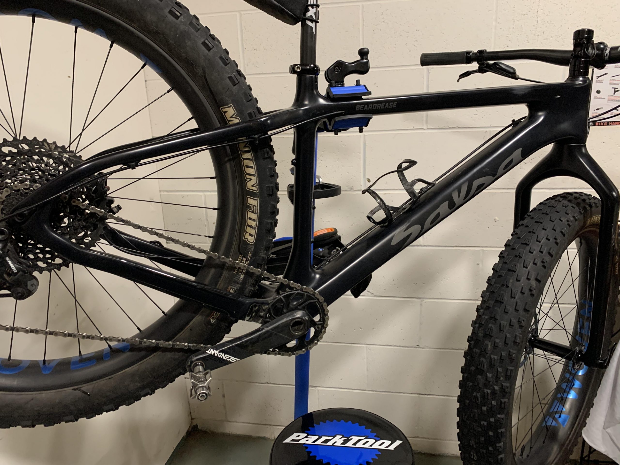Salsa Beargrease: A black fat bike is mounted on a repair stand, showcasing its frame, drivetrain, and large tires. The bike is equipped with a rear cassette and chain, while a blue and black Park Tool mat is visible beneath it. The background features a plain white wall, adding to the focus on the bike.