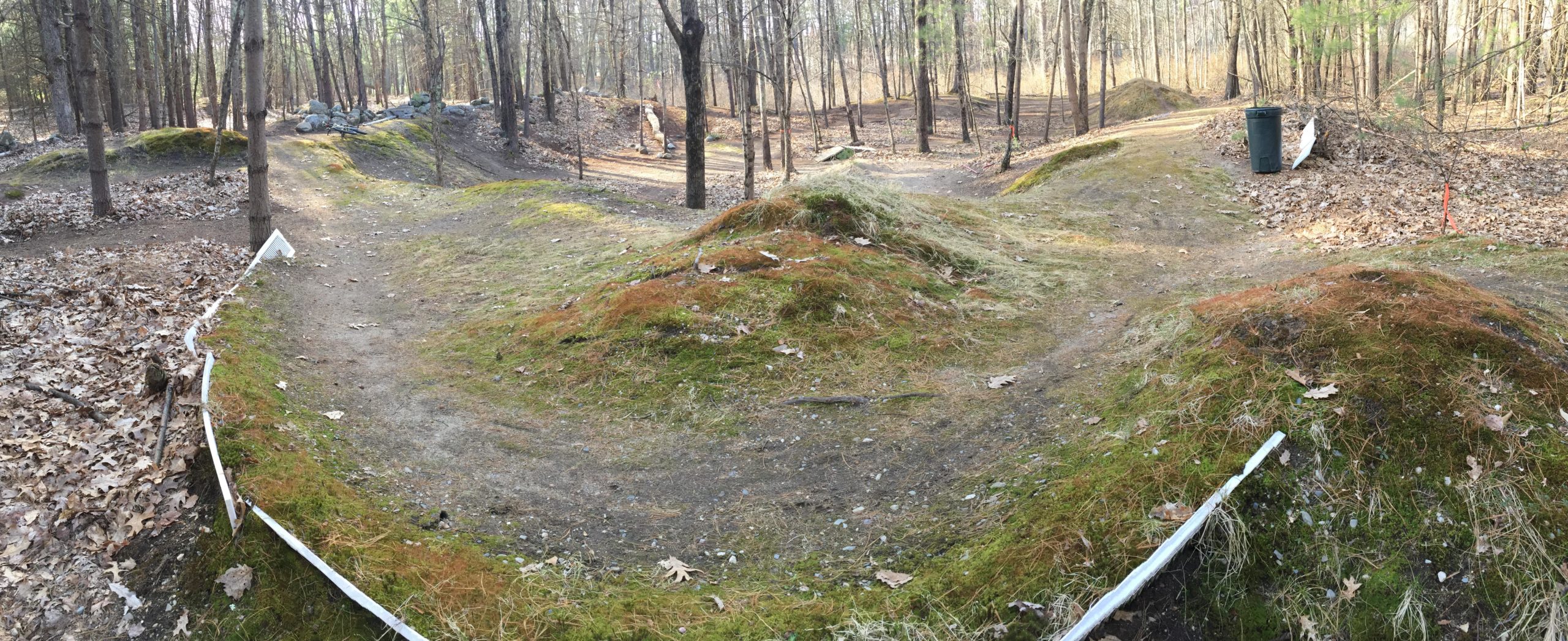 A panoramic view of a bike trail in a wooded area, featuring dirt paths winding around small hills covered in moss and fallen leaves. Trees surround the trail, with patches of sunlight filtering through the branches. A trash bin and a white sign can be seen in the background. Hitchinpost Greens mountain bike trail.