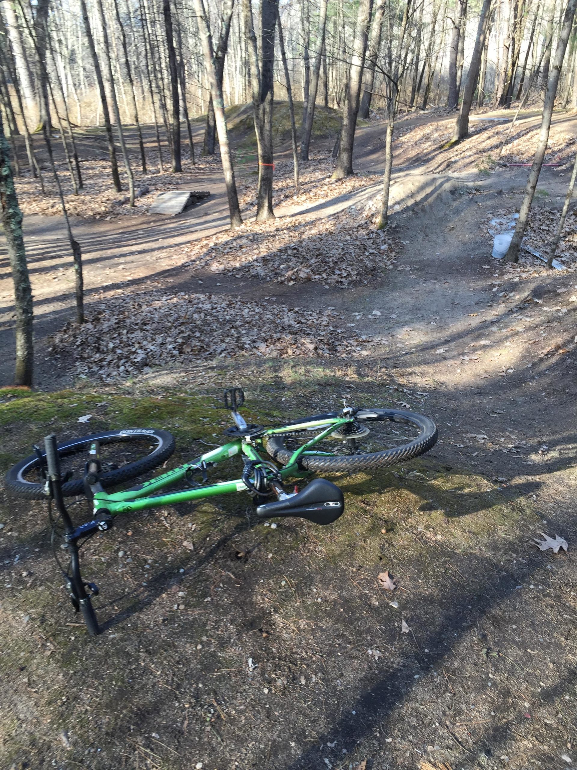 A green mountain bike laying on its side on a dirt path in a wooded area. Surrounding the bike are trees with bare branches and scattered autumn leaves on the ground. In the background, a small jump and winding trails are visible. Hitchinpost Greens mountain bike trail.